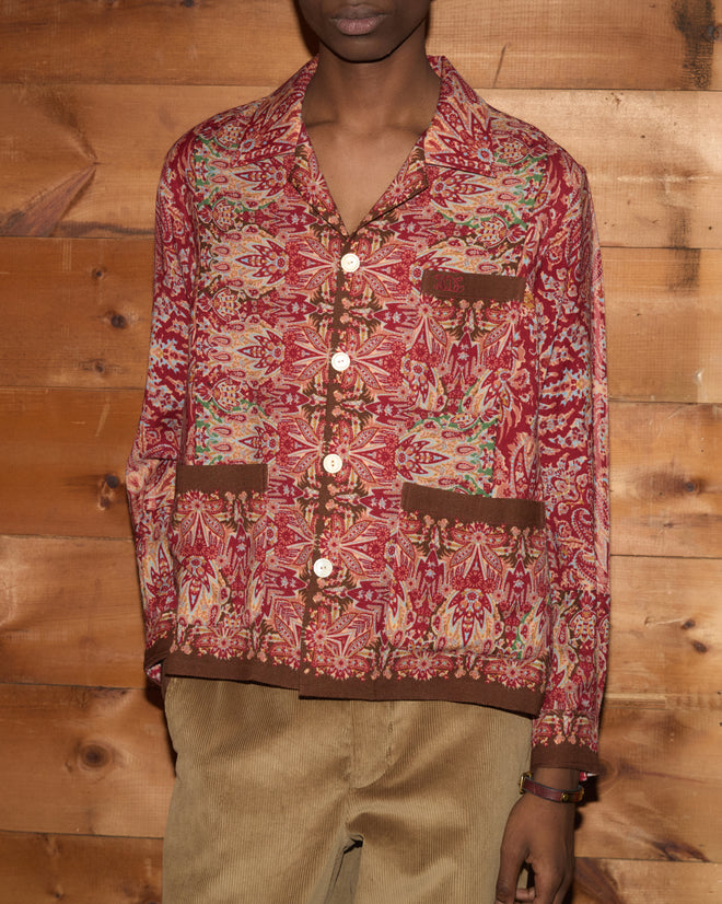 Person wearing the Arrowleaf Long Sleeve Shirt in a red paisley print and tan corduroy pants, standing against a wooden wall.
