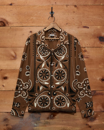 Bandana Print Long Sleeve Shirt featuring a brown paisley and floral design inspired by historic bandana patterns, button-up style, hanging on a hook against a wood wall.