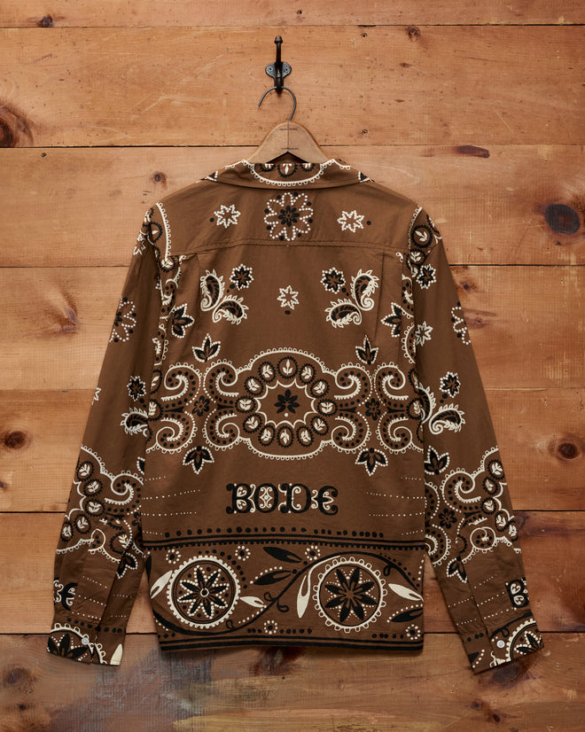 A Bandana Print Long Sleeve Shirt in brown cotton, featuring a white historic bandana pattern, hangs on a hook against a wooden wall.