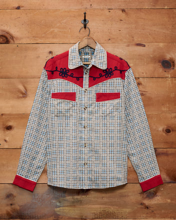 The River City Western Shirt, showcasing a plaid design with striking red yoke, cuffs, and pockets and embellished with black floral embroidery, hangs on a wooden wall.