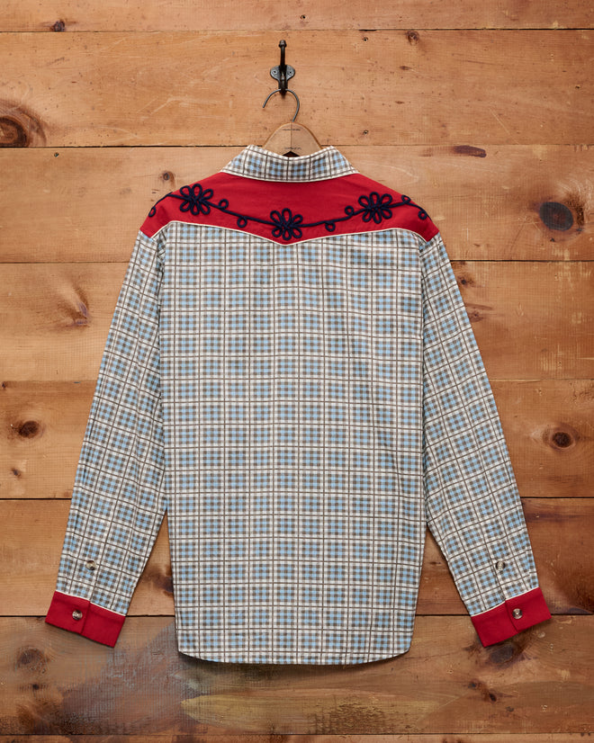 River City Western Shirt—a plaid long-sleeve cotton shirt with a red Western yoke and cuffs, finished with black floral trim—hanging on a wooden wall.