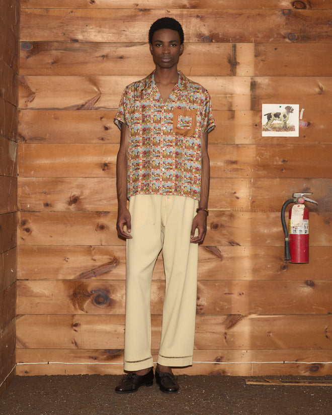 A person stands indoors by a wooden wall near a fire extinguisher, wearing the Cowboy Comic Short Sleeve Shirt paired with cream pants.