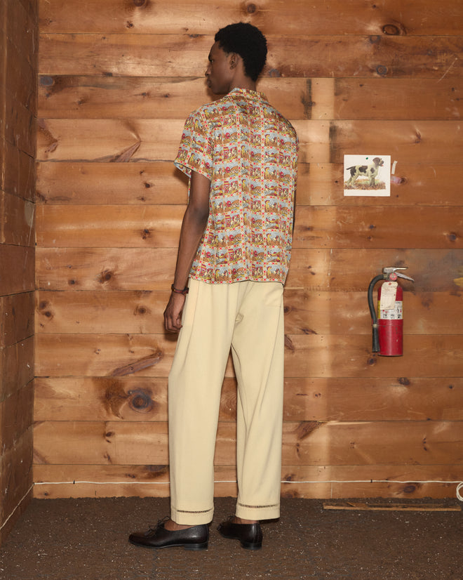 Wearing the Cowboy Comic Short Sleeve Shirt with its bold, colorful print and beige pants, a person stands facing a wooden wall decorated with a fire extinguisher and a dog photo, embodying the vibrant charisma of classic Western comics.