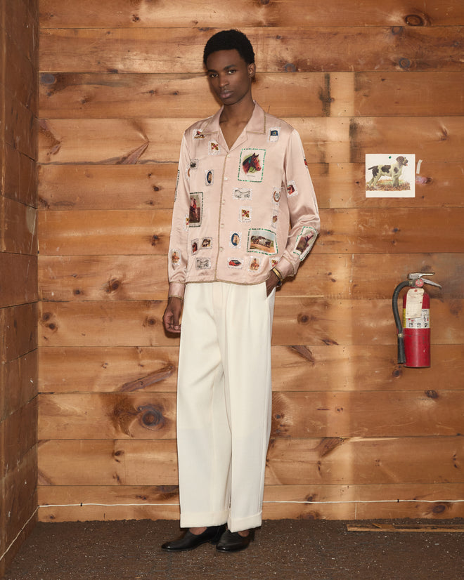 A person wearing the Equestrian Patch Long Sleeve Shirt featuring pink reproduction appliqué with printed patches reminiscent of collectible fabric swatches, paired with white pants, stands in a wooden room near a fire extinguisher.