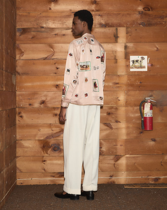 Wearing the Equestrian Patch Long Sleeve Shirt, featuring pink patterns reminiscent of vintage tobacco silks and collectible fabric swatches, and paired with white pants, a person stands by a fire extinguisher against a wooden wall.