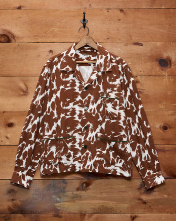 The Flannel Cow Print Long Sleeve Shirt, featuring a brown and white cow pattern and button details, is displayed hanging on a wooden wall with visible wood texture.