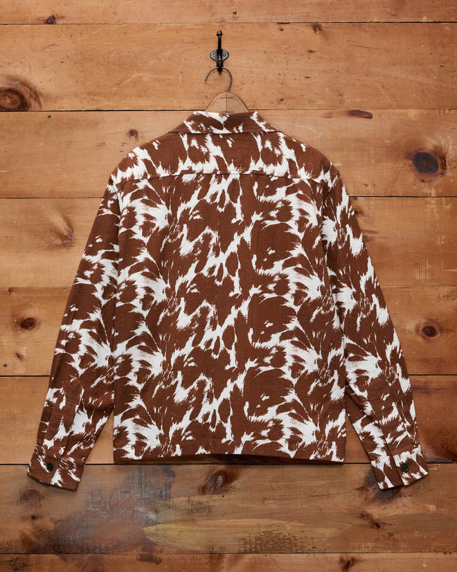 The Flannel Cow Print Long Sleeve Shirt features a brown and white cow print pattern, crafted from soft 100% cotton flannel. Hanging on a wooden wall by a metal hook, it’s the perfect Western shirt for children.