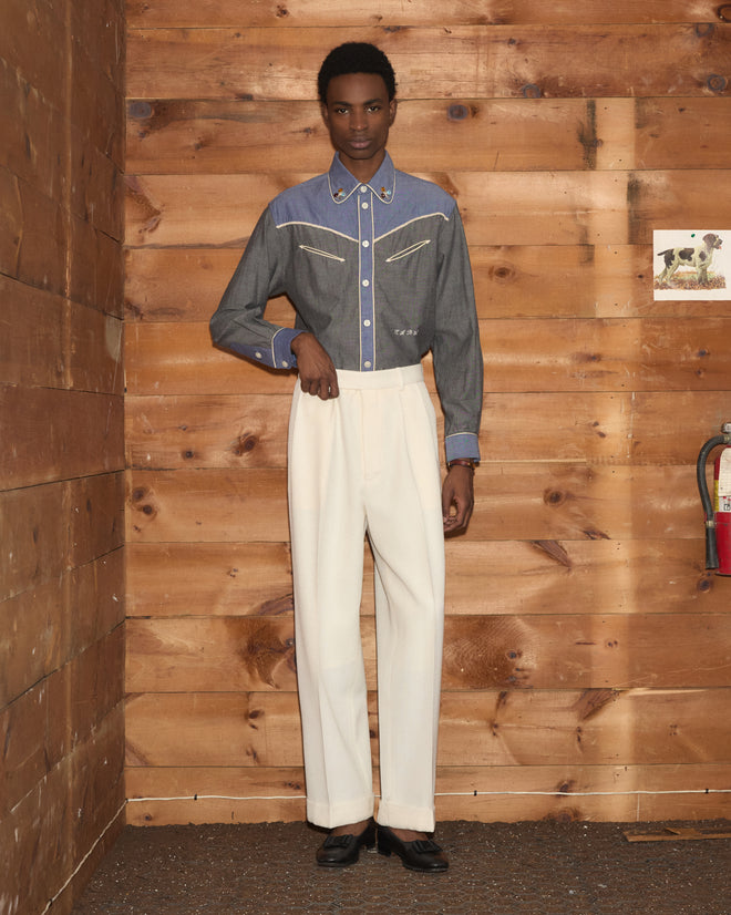 A person poses indoors by wooden walls wearing the Chambray Braid Western Shirt with a classic western yoke, paired with ivory trousers and black shoes.