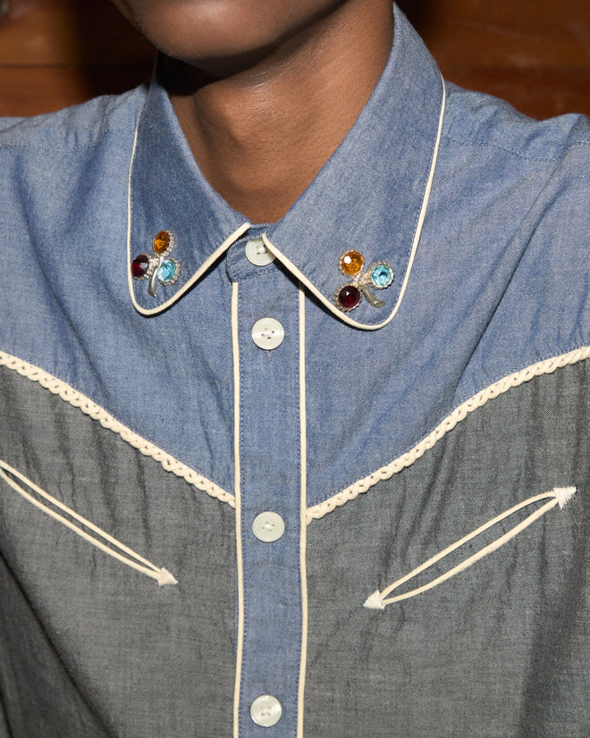 A close-up of the Chambray Braid Western Shirt, featuring a blue chambray fabric, rhinestone accents on the collar, white piping, and a classic western yoke.