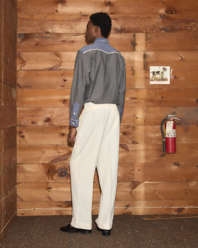 Wearing the Chambray Braid Western Shirt styled with white pants, a person stands facing a wooden wall featuring a fire extinguisher and a small picture.