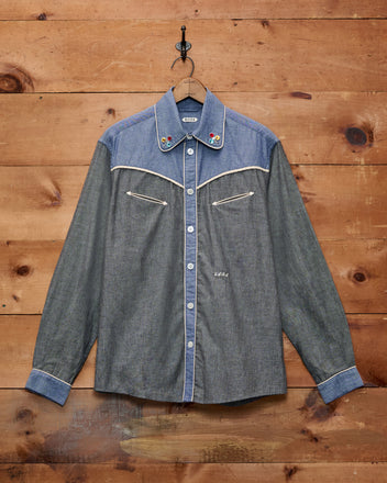 Chambray Braid Western Shirt in gray, featuring embroidery, smile pockets, white piping, and a classic Western yoke, displayed hanging on a wooden wall.