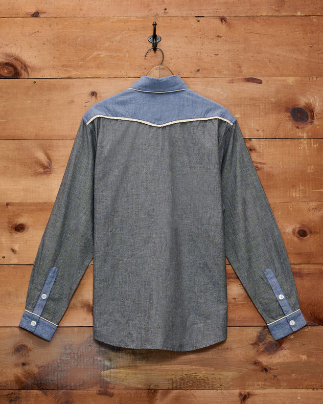 The Chambray Braid Western Shirt, featuring long sleeves and a Western yoke, hangs on a hook against a wooden wall, displayed from the back with distinctive white trim details.