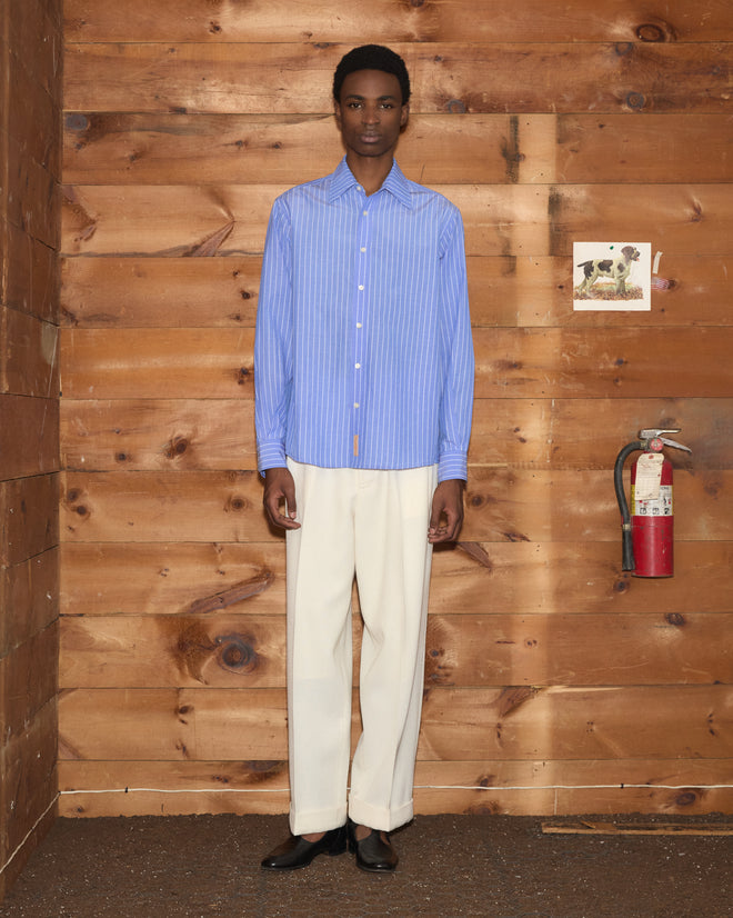 A person wearing the Bluebell Long Sleeve Shirt made from 100% cotton and cream pants stands indoors against a wooden wall near a fire extinguisher.