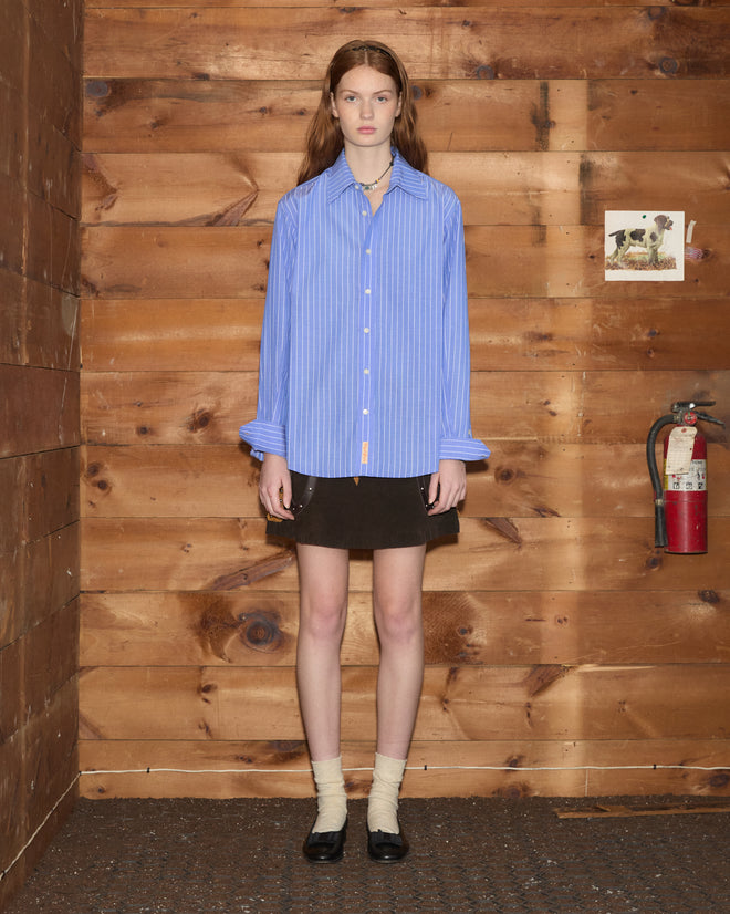 A woman wearing the Bluebell Long Sleeve Shirt and a black skirt stands in a wood-paneled room next to a fire extinguisher.