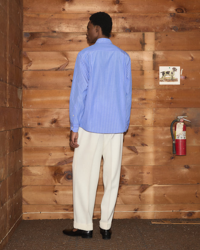 A person in the Bluebell Long Sleeve Shirt—a vintage shirting-inspired, blue striped 100% cotton shirt—and white pants stands facing a wooden wall with a fire extinguisher.