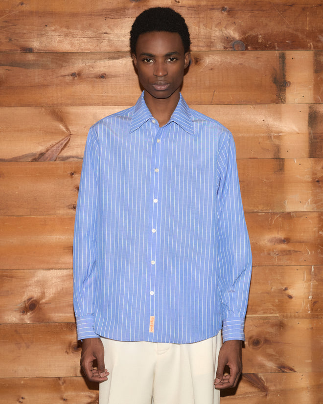 Person wearing the Bluebell Long Sleeve Shirt, made from 100% cotton with a vintage striped design, paired with cream pants and standing in front of a wooden wall.