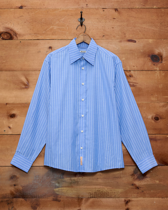 The Bluebell Long Sleeve Shirt, made from 100% cotton in a blue-and-white pinstripe design, hangs on a hook against a wooden wall.