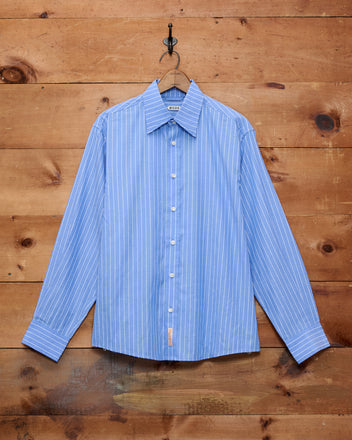 The Bluebell Long Sleeve Shirt, made from 100% cotton in a blue-and-white pinstripe design, hangs on a hook against a wooden wall.