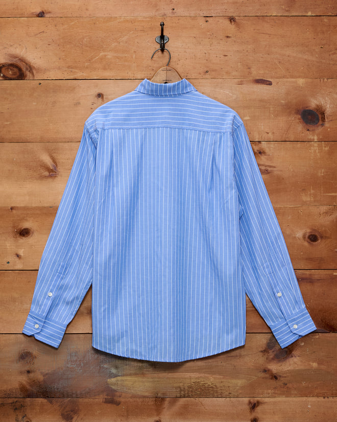 The Bluebell Long Sleeve Shirt, featuring a collared design with white-accented blue stripes, is crafted from premium 100% cotton shirting and hangs on a wooden wall.