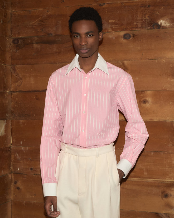 A person wearing the Pickfair Shirt, featuring pink stripes and a contrasting white collar, paired with cream pants, stands in front of a wooden wall looking at the camera.