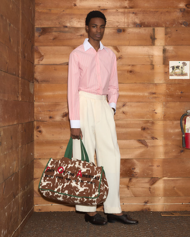 A person wearing the Pickfair Shirt in pink, featuring a contrasting white collar, pairs it with white pants while standing indoors and holding a large camo-print bag with green handles.
