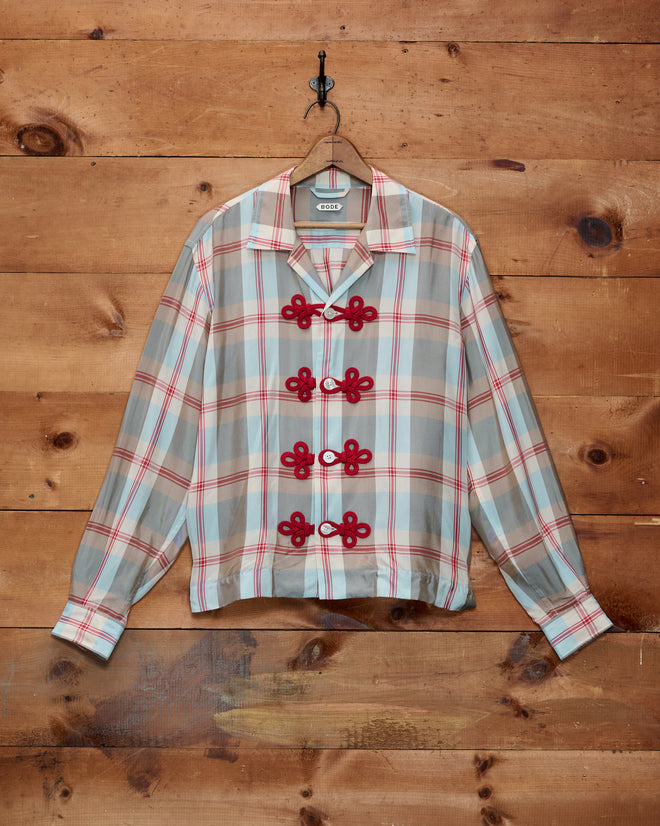 Canyon Trillium Shirt—a classic red and blue plaid button-up featuring elegant decorative frog closures, hanging on a hook against a wooden wall.