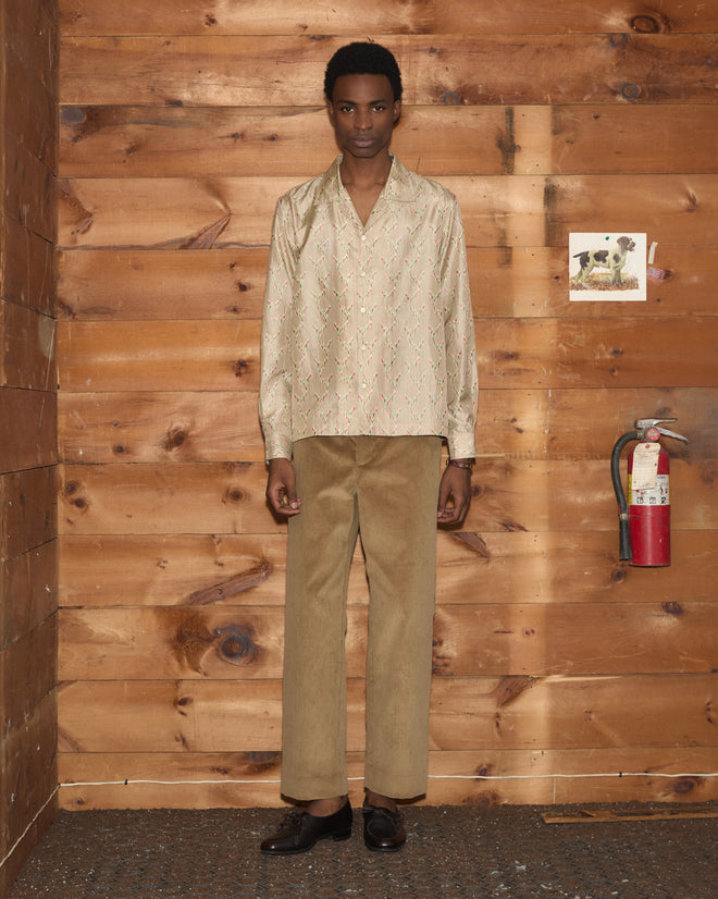 A person wearing the Sagebrush Long Sleeve, a cream patterned silk shirt with a boxy fit, and tan pants stands in a wooden room near a fire extinguisher on the wall.
