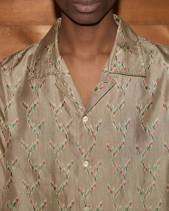 A person wearing the Sagebrush Long Sleeve—a tan patterned, collared button-up shirt with a boxy fit—stands in front of a wooden background.