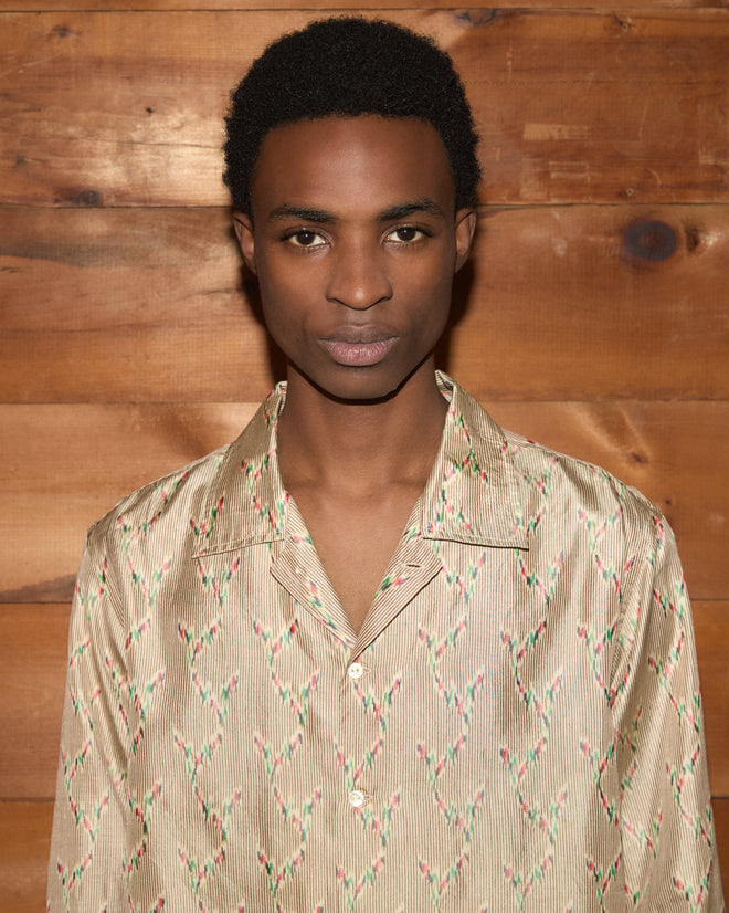 A person wearing the Sagebrush Long Sleeve—a boxy fit, patterned beige shirt—stands in front of a wooden wall, facing the camera.
