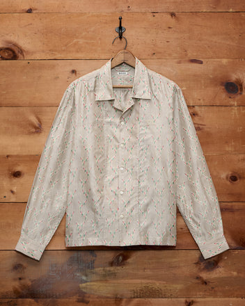Sagebrush Long Sleeve—beige silk shirt with a subtle geometric pattern, boxy fit, and button-up front—hanging on a hook against a wooden wall.