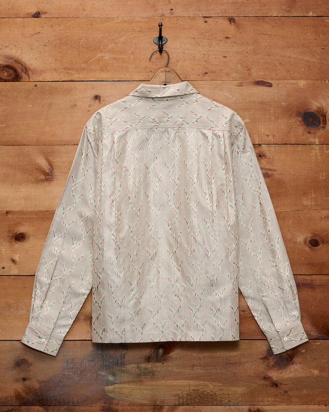 A Sagebrush Long Sleeve shirt, featuring a beige patterned silk fabric and boxy fit, hangs on a black hook against a wooden wall.