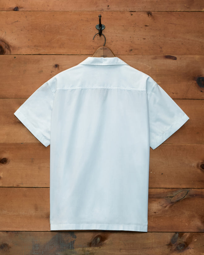 Back view of a white, 100% cotton poplin Clover Charm Short Sleeve Shirt on a hanger against a wood wall.