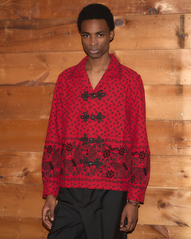 A person stands in front of a wooden wall, looking at the camera while wearing the Beaded Kerchief Long Sleeve Shirt featuring a red and black pattern with black beadwork.