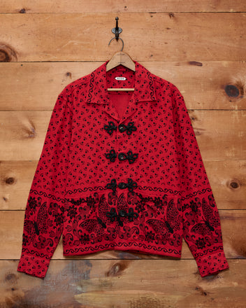 Beaded Kerchief Long Sleeve Shirt featuring red patterns, black beadwork, and distinctive frog closures hangs on a wooden wall.