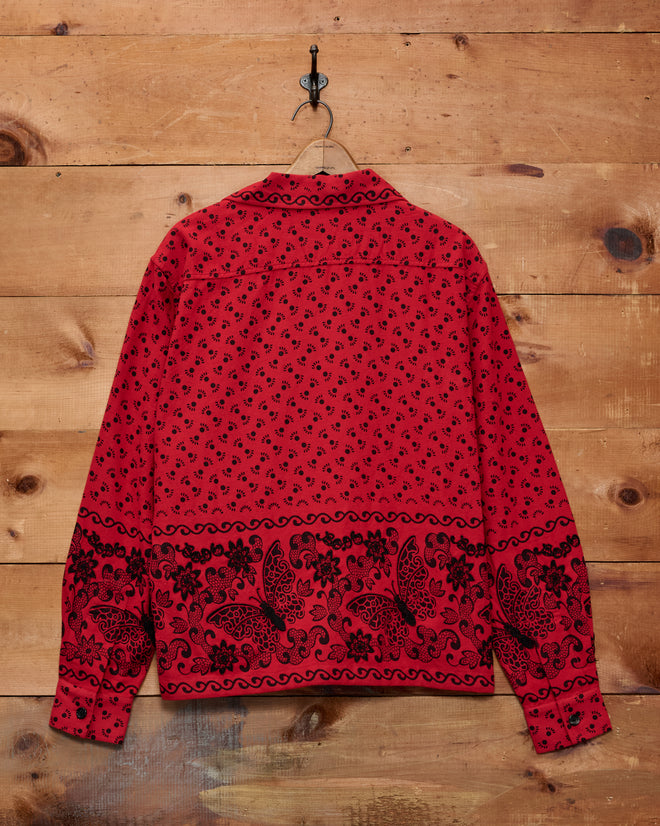 A Beaded Kerchief Long Sleeve Shirt featuring black floral and paisley patterns hangs on a hook against a wooden wall, its red fabric accented with subtle black beadwork.