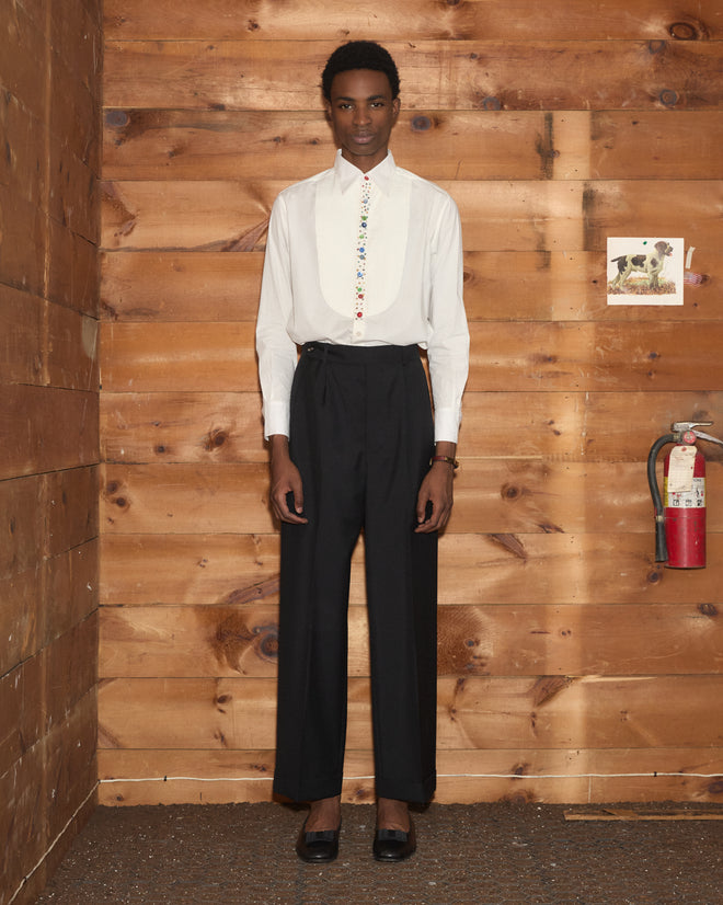 Person in a Button Jar Tuxedo Shirt with a pleated bib and black pants stands indoors against a wooden wall next to a fire extinguisher.