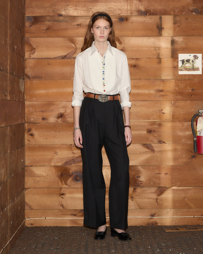 A woman stands against a wooden wall next to a fire extinguisher, wearing the Button Jar Tuxedo Shirt—a white shirt with a pleated bib, crafted from 100% cotton for lasting comfort and style—paired with black pants and a headband.