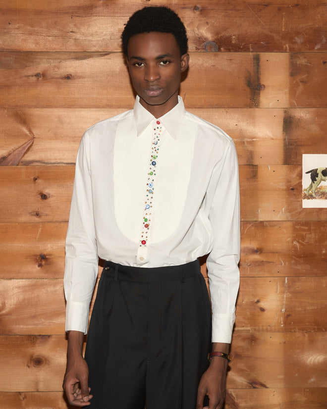Wearing the Button Jar Tuxedo Shirt—a white tuxedo shirt with a pleated bib and colorful buttons—paired with black pants, a person stands before a wooden wall.