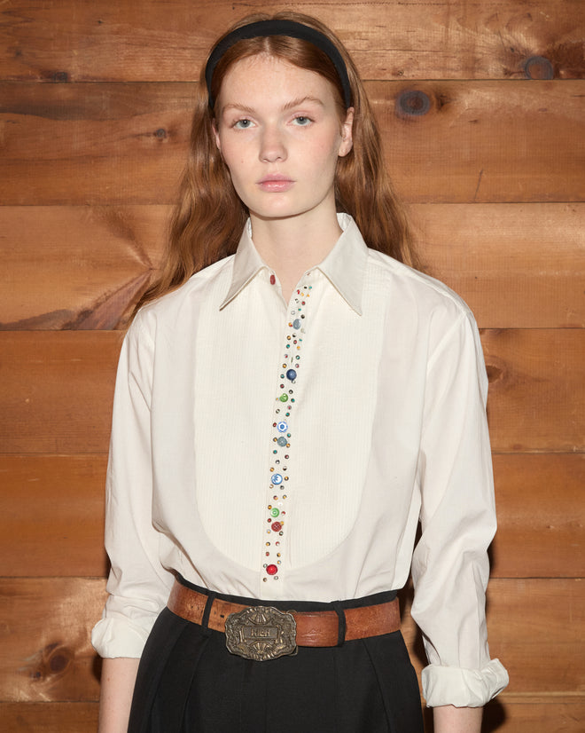 A woman stands against wood paneling wearing the Button Jar Tuxedo Shirt—crafted from 100% cotton for comfortable elegance—with a pleated bib and colorful buttons, paired with black pants and a brown belt.