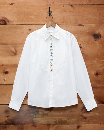 The Button Jar Tuxedo Shirt, crafted from 100% cotton, features a pleated textured bib and multicolored buttons, displayed against a wooden wall.