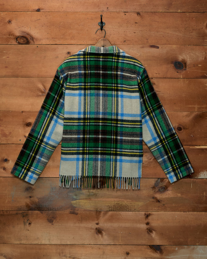 Mr. Checkers Overshirt in green, blue, and white plaid with a fringed hem, hanging on a wooden wall.