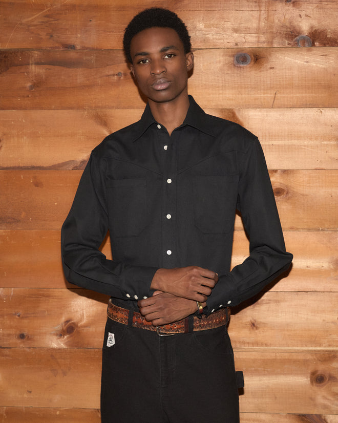 A man wearing the Faille Western Shirt, featuring a classic black color and Western yoke, stands in front of a wooden wall with his arms crossed.