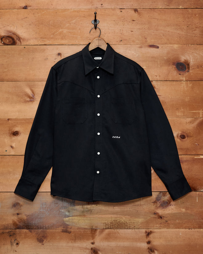 The Faille Western Shirt—a black button-up with two chest pockets and a classic Western yoke—hangs on a wooden wall from a metal hook.