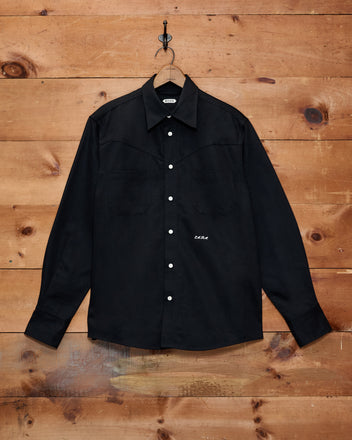 The Faille Western Shirt—a black button-up with two chest pockets and a classic Western yoke—hangs on a wooden wall from a metal hook.