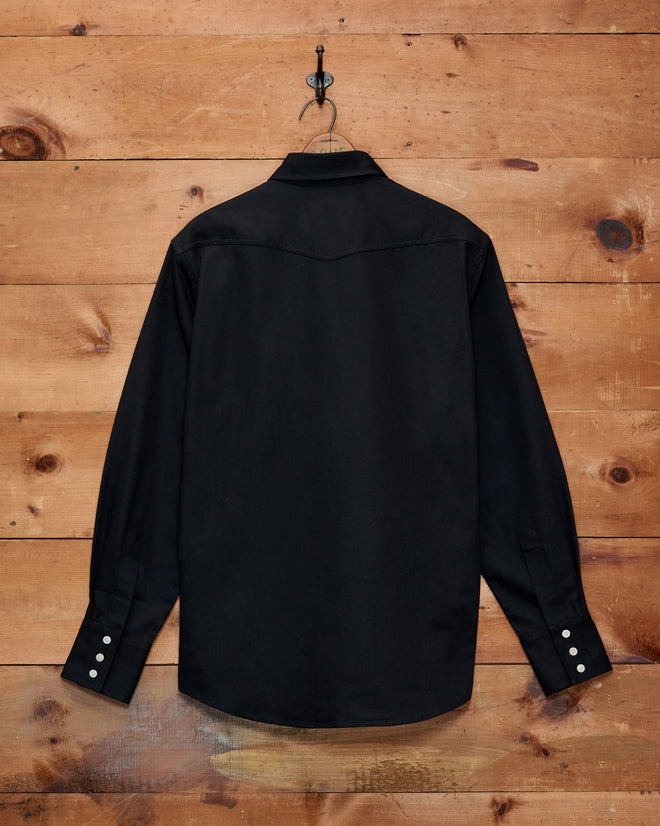 A Faille Western Shirt in black with long sleeves hangs on a hook against a wooden wall, displayed from the back.