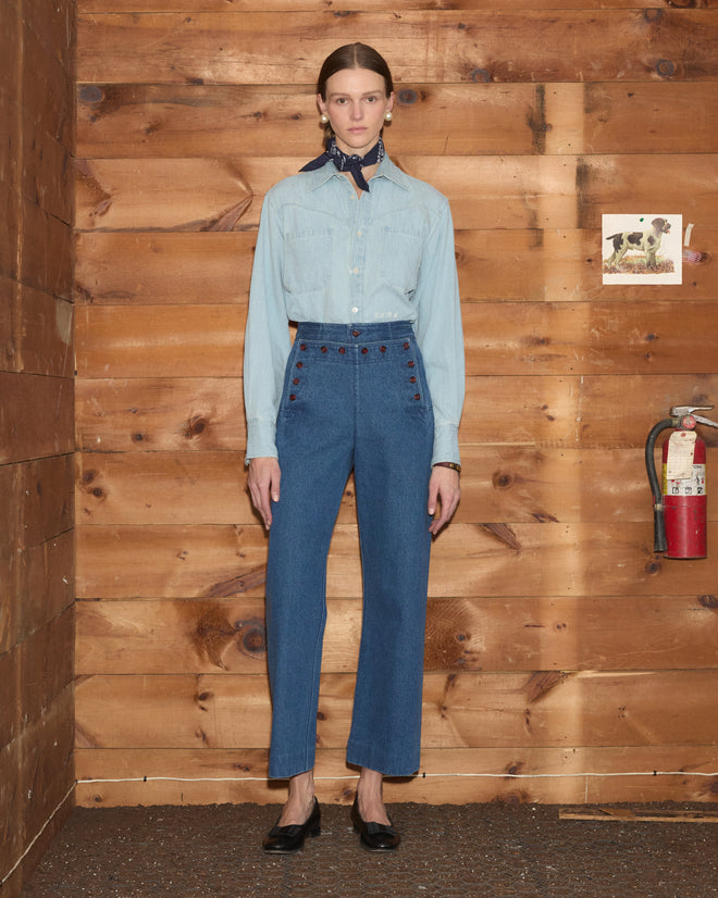 A woman in a blue 100% cotton denim shirt and midweight navy Denim Sailor Trousers stands indoors against a wooden wall, near a fire extinguisher.