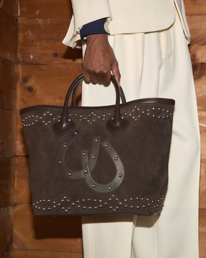 A person in cream pants holds the Studded Horseshoe Tote, a brown suede bag accented with studs and a leather horseshoe appliqué, crafted from delicate cow suede for a refined finish.