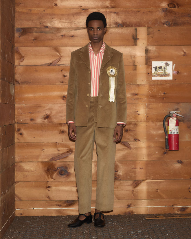 A person in a brown corduroy suit with Corduroy Side-Buckle Trousers, a striped shirt, and a rosette lapel pin.