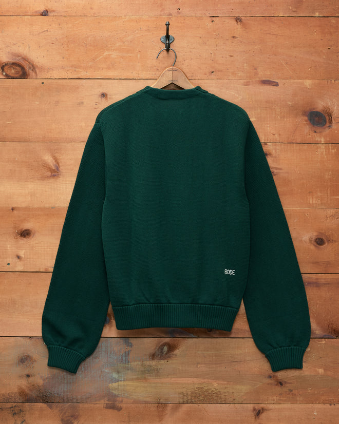 A dark green Double-Breasted Cardigan hangs on a wooden wall, displayed from the back. “BODE” is embroidered near the waistband, highlighting this unique piece.