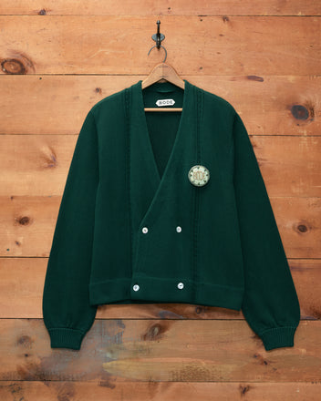 Double-Breasted Cardigan - Dark Green with double-breasted buttons and a pin, displayed on a wooden wall.